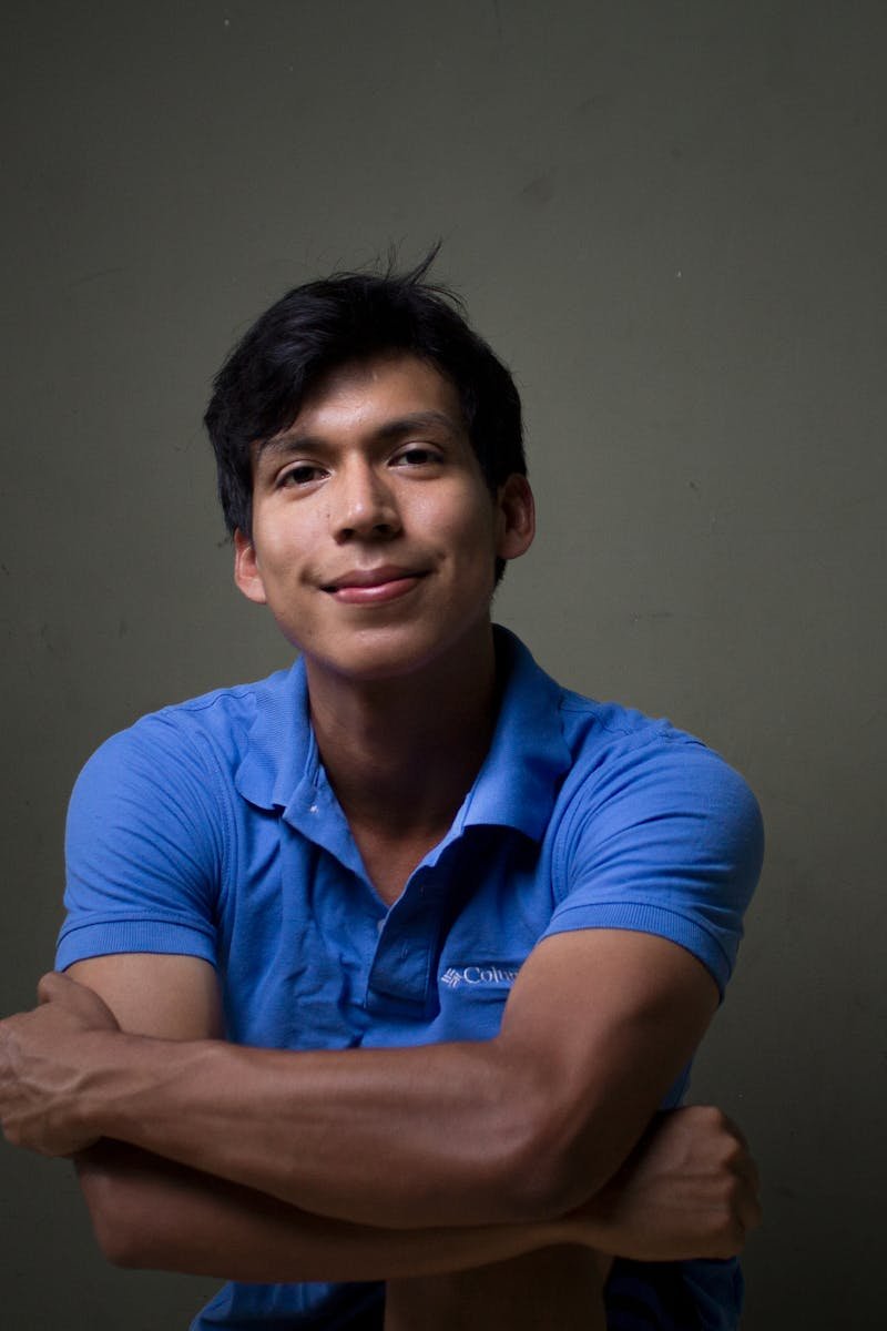 Young Hispanic man smiling confidently in a blue shirt against a dark background.